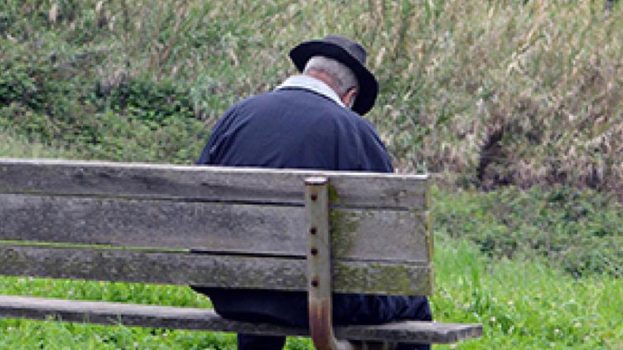 An elderly man, head bowed, sits alone on a bench. 