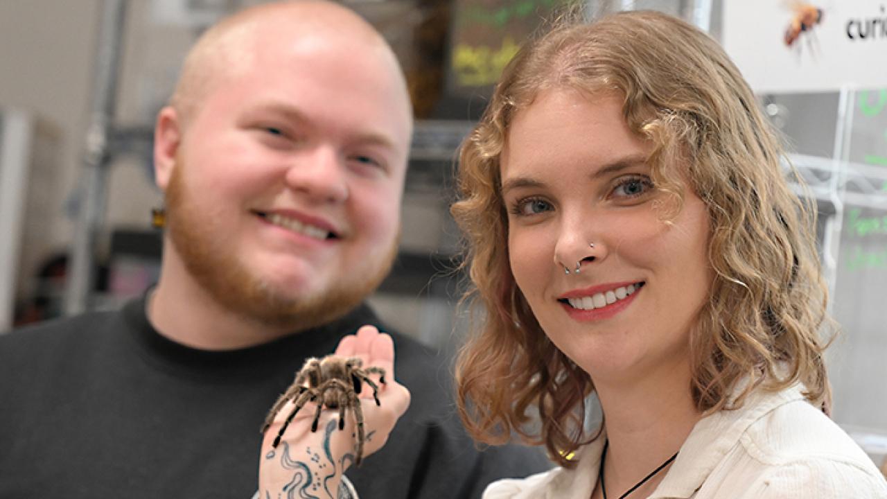 Doctoral candidate Emma Jochim holds a tarantula while smiling next to Felix Duley in a lab setting.