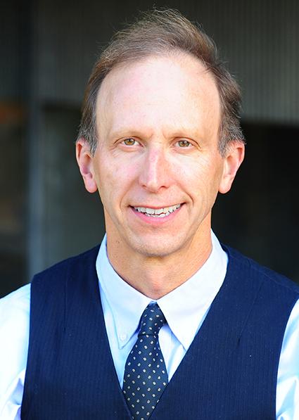 Smiling middle-aged man headshot in navy vest, white shirt and polka-dot tie, neutral background