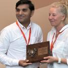 Two people in white shirts with orange lanyards holding a wooden award plaque, smiling