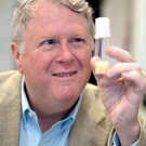 A man in a lab coat holds a small test tube with a smile, surrounded by laboratory equipment.